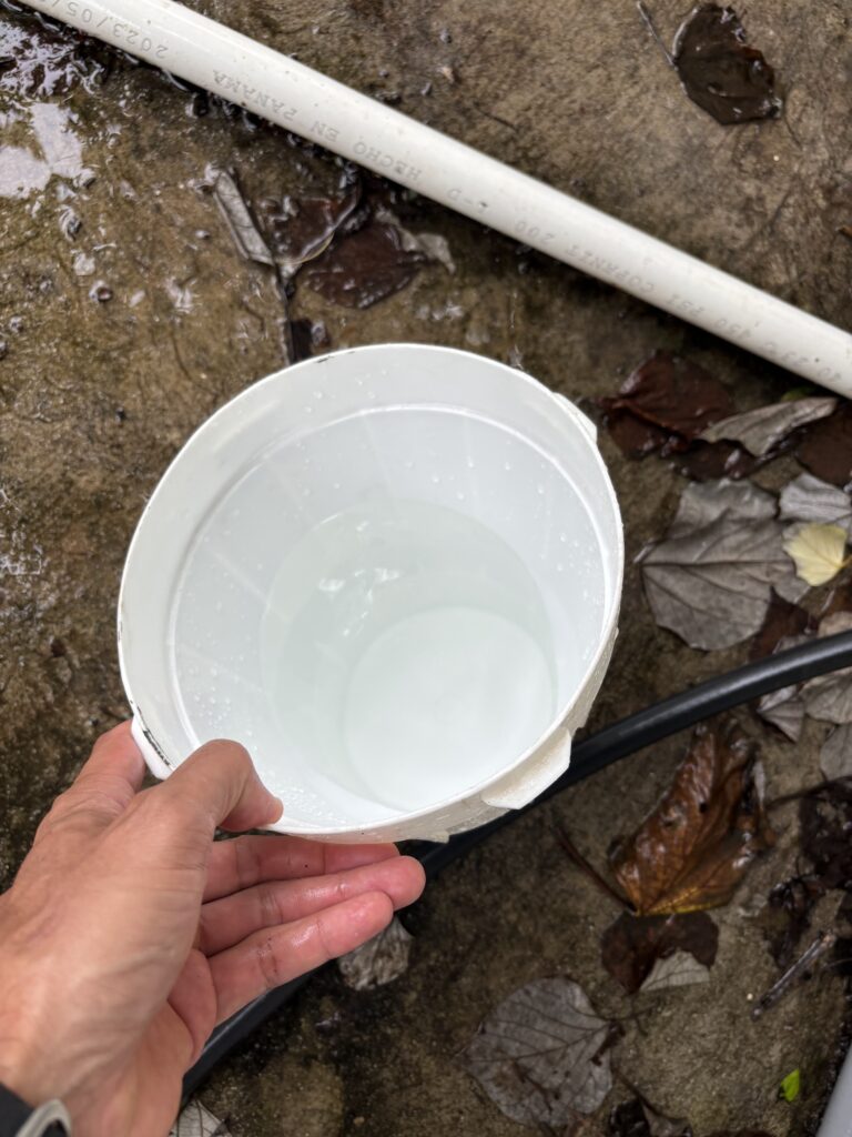 Una persona sostiene un recipiente con una muestra de agua ligeramente turbia junto a tubería PVC, sobre el terreno húmedo de una instalación de pozo.