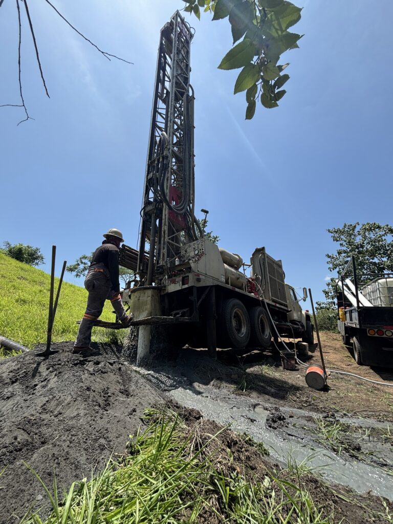 Perforadora montada en camión trabajando bajo cielo despejado; un operario con casco maneja el equipo mientras sale lodo de perforación del terreno.