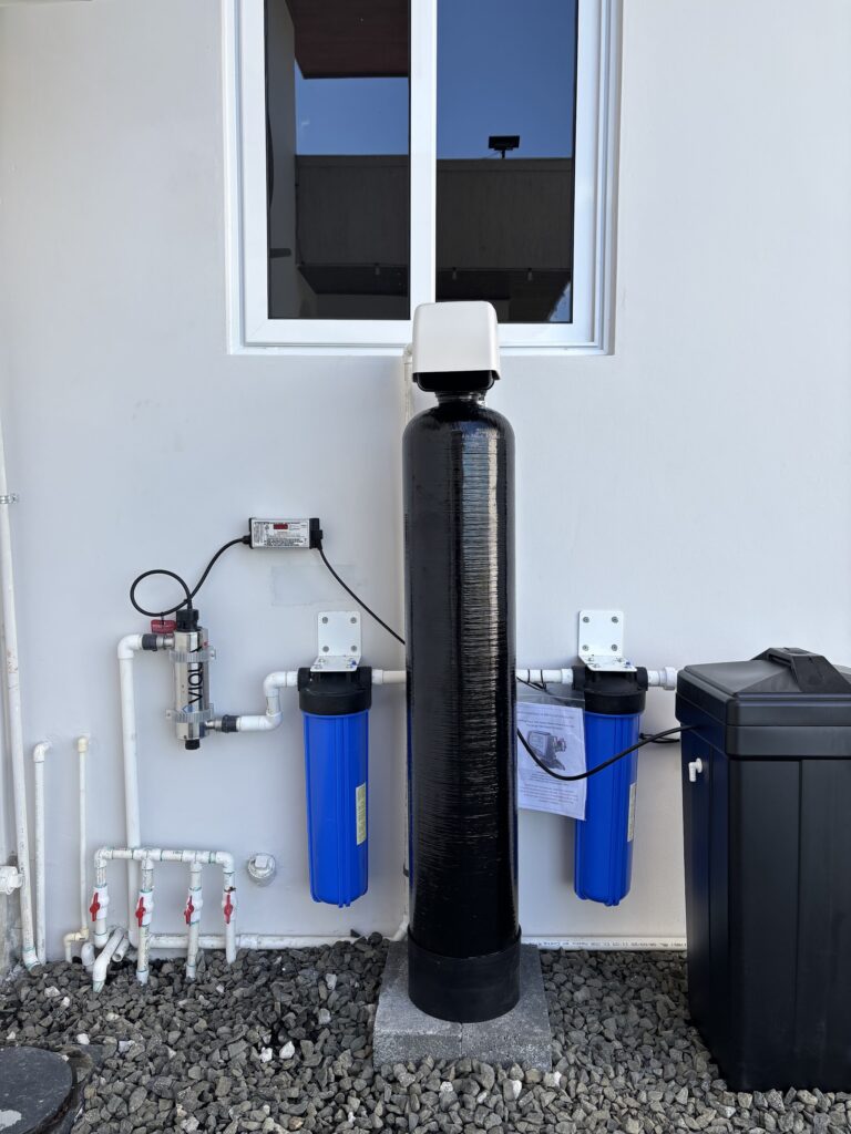 Sistema exterior sobre grava seca: ablandador negro, luz UV Viqua, dos filtros azules y tanque de carbón, montados en la pared de una vivienda.